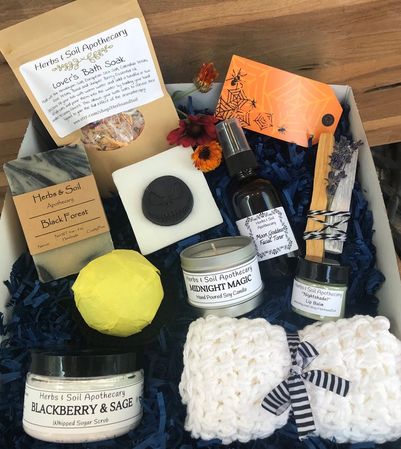 May include: A Halloween-themed gift box filled with bath and body products from Herbs & Soil Apothecary. The box includes a black forest soap, a blackberry and sage whipped sugar scrub, a midnight magic soy candle, a moon goddess facial toner, a nightshade lip balm, a bath bomb, a crocheted washcloth, and a wooden stick of incense.