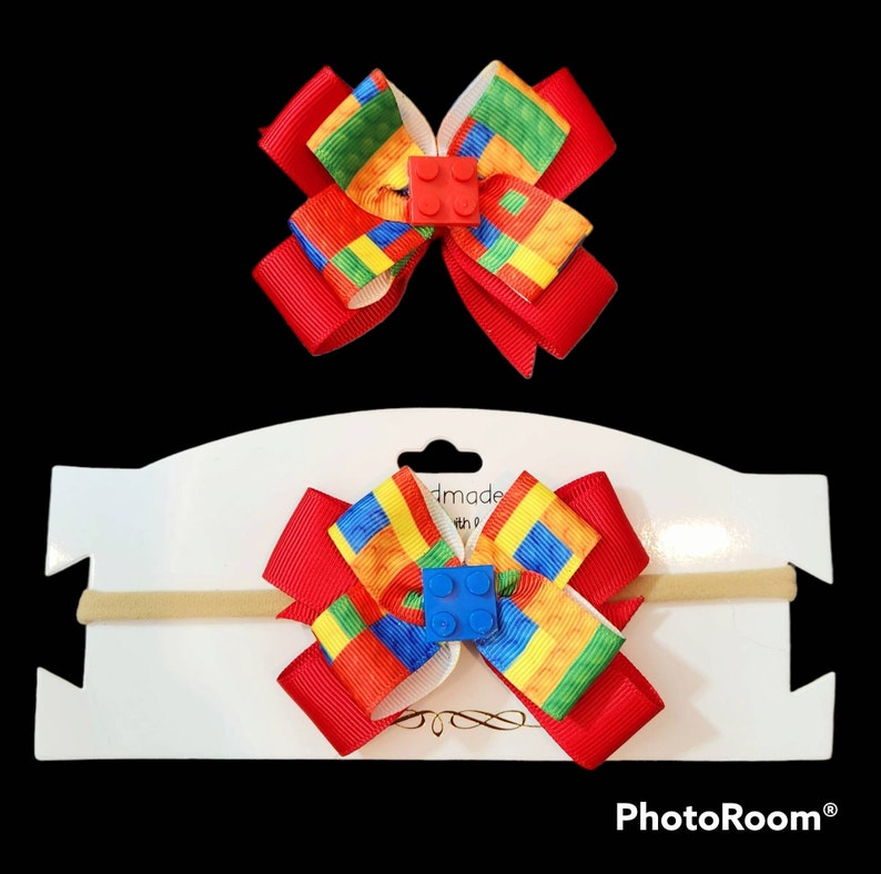 May include: A red and white bow with a Lego block design and a blue Lego block in the center. The bow is attached to a nude elastic headband.