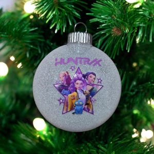May include: Glittery silver Christmas ornament with the word "HUNTRAX" in purple. The ornament features a graphic of three women and a blue cat inside a purple star. The ornament hangs on a green Christmas tree.