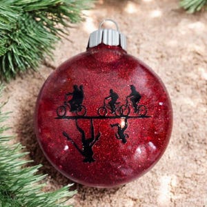 May include: Red glitter Christmas ornament with a black silhouette design of three people riding bicycles above a tree and a person hanging upside down. The ornament has a silver metal cap and is surrounded by pine branches.