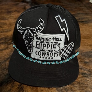 May include: Black trucker hat with a turquoise beaded trim. The hat features several patches, including a skull, a lightning bolt, a checkered heart, and a patch that reads "Raising Hell with the Hippies and the Cowboys".