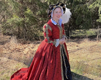 May include: A woman wearing a red and black dress with gold trim and a black hat with gold accents. She is standing in a grassy area with trees in the background.