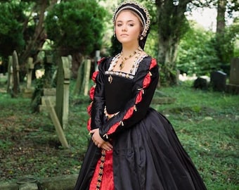 May include: A black and red long-sleeved gown featuring a square neckline, fitted bodice, and full skirt. The sleeves have red fabric puffs, and the front panel is red with gold trim. A beaded headpiece and gold necklace complete the ensemble.