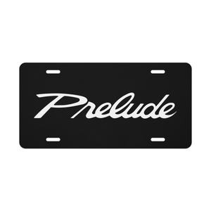 May include: A rectangular black license plate featuring the word Prelude written in a white stylized script font across the center. The plate includes four horizontal mounting slots, two positioned along the top edge and two along the bottom edge.