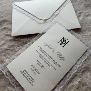 May include: Ivory wedding invitation set with a matching envelope. The invitation features delicate lace trim and a pearl accent on the envelope. The invitation includes the names "Label & Mustafa" and event details.