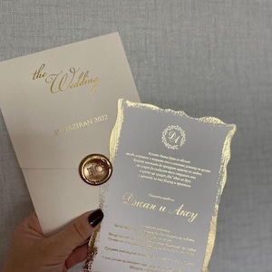 Può includere: Invito di nozze color avorio con testo e dettagli in lamina d'oro. L'invito presenta le parole "the Wedding" e "15 HAZIRAN 2022". Un sigillo di cera dorata e un bordo decorativo incorniciano il testo dell'invito.