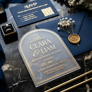 May include: An arched frosted acrylic wedding invitation with gold lettering reading Clara & Liam, accompanied by a navy blue RSVP card with a QR code, a navy envelope with a gold wax seal, and a navy velvet ring box containing two gold rings.