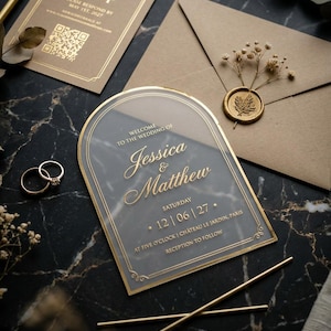 Könnte beinhalten: Eine bogenförmige, transparente Acryl-Hochzeitseinladung mit goldener Schrift, die Jessica & Matthew, 12 | 06 | 27 und Château Le Jardin, Paris zeigt. Enthält eine RSVP-Karte mit QR-Code, einen braunen Umschlag mit goldenem Wachssiegel und zwei Goldringe.