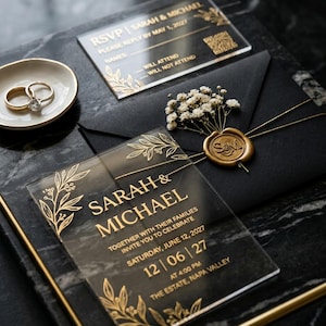 Op de afbeelding: Een elegante trouwuitnodigingenset met heldere acrylkaarten met gouden letters en bloemaccenten. Bevat een uitnodiging, een RSVP-kaart en een zwarte envelop verzegeld met een gouden wasstempel. Twee ringen liggen in een klein schaaltje.