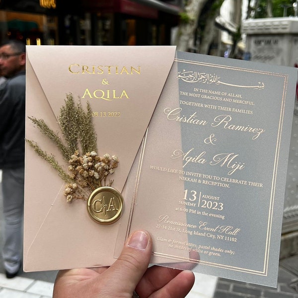 May include: A wedding invitation suite featuring a blush pink envelope with gold lettering and a sprig of dried flowers, sealed with a gold wax stamp. The invitation card is gray with gold text, including the names Cristian Ramirez & Agila Miji.