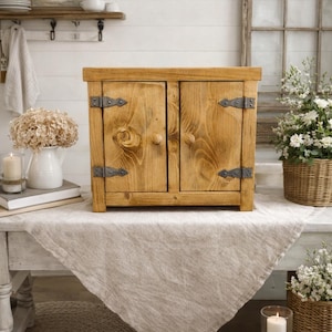 May include: A small, rustic wooden cabinet with two doors, featuring visible wood grain and dark metal hinges. The cabinet sits on a table covered with a neutral-coloured linen cloth, creating a farmhouse aesthetic.