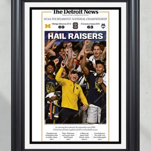 May include: Framed print of The Detroit News front page, celebrating the Michigan Wolverines' NCAA National Championship victory. The headline "HAIL RAISERS" is prominent, with a photo of jubilant fans. The score is displayed: Michigan 69, Connecticut 63.