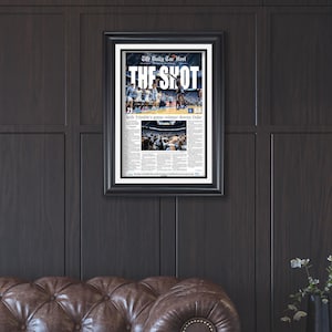 May include: Framed black and white newspaper print titled "The Shot" with a basketball game image. The headline reads "Seth Trimble's game-winner downs Duke." The print is displayed on a dark wood paneled wall.