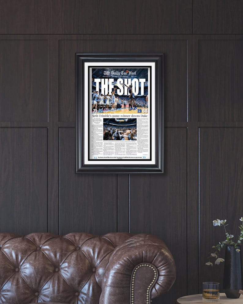 May include: Framed black and white newspaper print titled "The Shot" with a basketball game image. The headline reads "Seth Trimble's game-winner downs Duke." The print is displayed on a dark wood paneled wall.