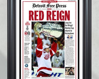 Op de afbeelding: Ingelijste voorpagina van de Detroit Free Press krant met de kop "RED REIGN". De afbeelding toont een hockeyspeler die de Stanley Cup vasthoudt. De krant bevat tekst over de overwinning van de Wings in de Stanley Cup.