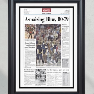 May include: Framed newspaper page with the headline "A-maizing Blue, 80-79" and sports photos. The black frame surrounds the white-bordered page. Text includes "Detroit Free Press" and "Robinson's OT free throws give U-M 1st NCAA title."