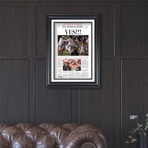 May include: Framed newspaper front page from The Boston Globe, with the headline "YES!!!" and images of a baseball game. The black frame contrasts with the dark wood paneling and a brown leather sofa.