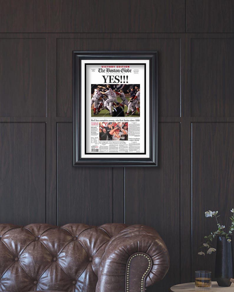 May include: Framed newspaper front page from The Boston Globe, with the headline "YES!!!" and images of a baseball game. The black frame contrasts with the dark wood paneling and a brown leather sofa.