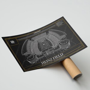 May include: A black and white architectural blueprint style print of Heinz Field, a football stadium in Pittsburgh, Pennsylvania. The print features the stadium's layout, including the field, seating, and surrounding buildings. The text "HEINZ FIELD" and "100 Art Rooney Avenue, Pittsburgh, Pennsylvania" is printed in white on a gold background.