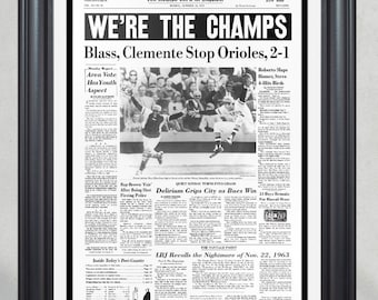 May include: A black and white newspaper clipping from the Pittsburgh Post-Gazette dated October 14, 1960. The headline reads "We're the Champs" and the sub-headline reads "Blass, Clemente Stop Orioles, 2-1".