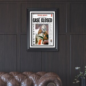 May include: A newspaper headline reads "CASE CLOSED" with the subheading "CANES CLOBBER HUSKERS, WIN FIFTH NATIONAL TITLE". The headline is in bold, black font with a red background. The newspaper is framed in black and is hanging on a dark wood wall.