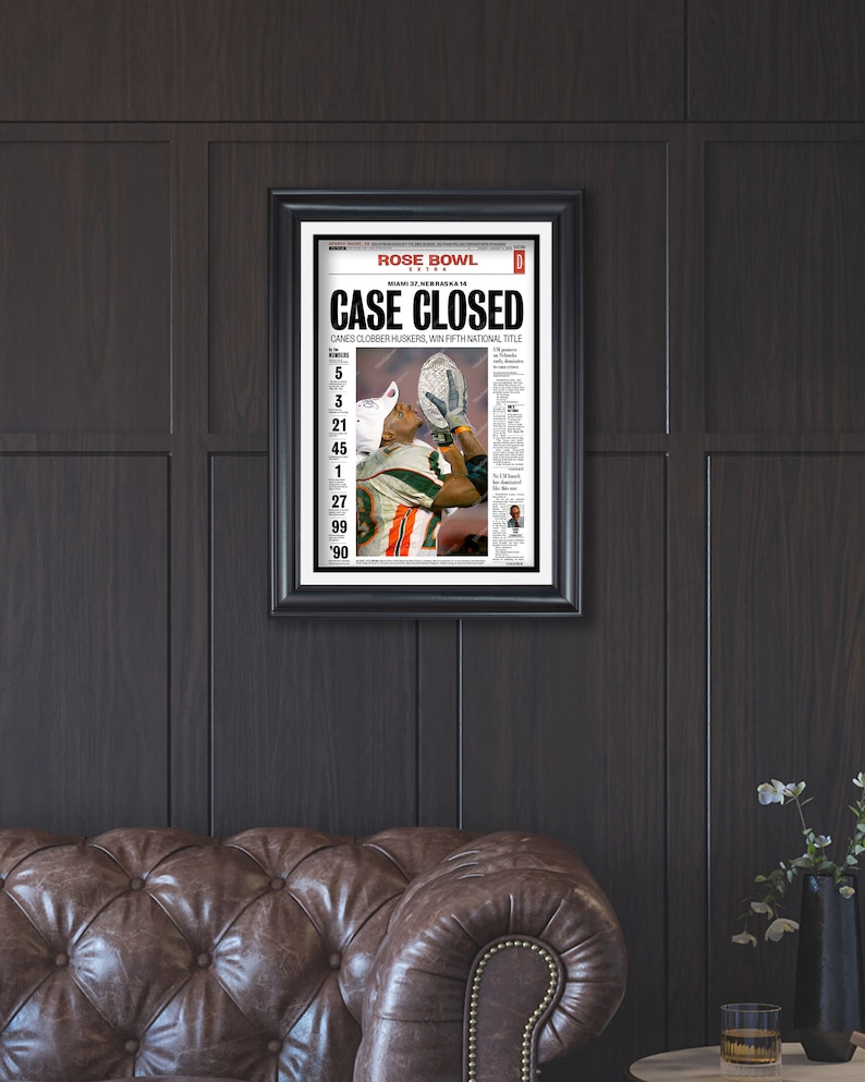 May include: A newspaper headline reads "CASE CLOSED" with the subheading "CANES CLOBBER HUSKERS, WIN FIFTH NATIONAL TITLE". The headline is in bold, black font with a red background. The newspaper is framed in black and is hanging on a dark wood wall.