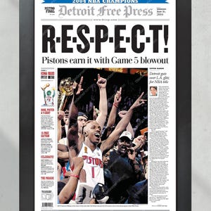 Może przedstawiać: Oprawiona strona tytułowa gazety Detroit Free Press, świętująca mistrz&oacute;w NBA 2004. Nagł&oacute;wek brzmi "R-E-S-P-E-C-T!" ze zdjęciem zespołu Pistons świętującego zwycięstwo. Rama jest czarna.
