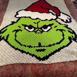 May include: A large, handmade crochet blanket featuring the Grinch's face. The Grinch is depicted in green with black outlines, white eyes, and a red Santa hat. The blanket has a neutral-colored background.