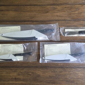 May include: Several kitchen knives, including a cleaver and a serrated knife, are packaged in clear plastic. The knives have black handles and are protected by white blade guards. The knives are on a wooden surface.