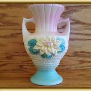 May include: A small, pink and blue ceramic vase with a white water lily design. The vase has two handles and a textured base.