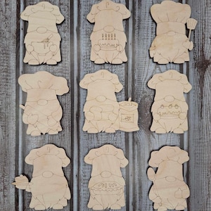 May include: Nine unfinished wooden gnome ornaments with different baking-themed designs. Each gnome is wearing a chef's hat and has a beard. The designs include a gnome holding a bowl with a whisk, a gnome holding a cake with candles, a gnome holding a spatula, a gnome holding a rolling pin, a gnome holding a bag of flour, a gnome holding a cake with strawberries, a gnome holding a plate with a pancake, a gnome holding a pie, and a gnome holding a spoon.