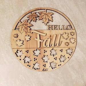 May include: A round wooden sign with the words "Hello Fall" in a decorative font. The sign is decorated with cut-out leaves and berries.