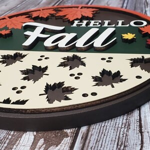 May include: A round wooden sign featuring the words "Hello Fall" in white lettering. The sign is decorated with autumn leaves in shades of red, orange, and yellow. The bottom half of the sign is cream coloured with a pattern of brown leaves.