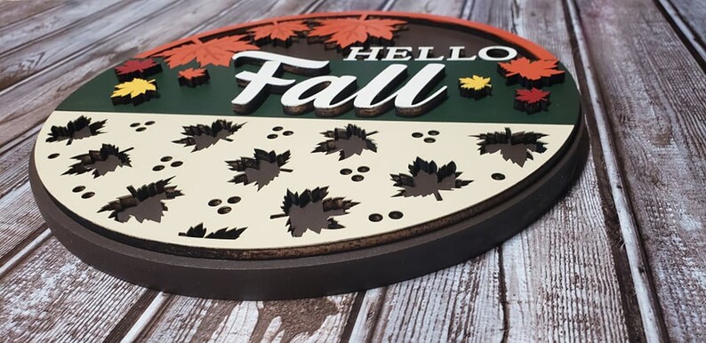 May include: A round wooden sign featuring the words "Hello Fall" in white lettering. The sign is decorated with autumn leaves in shades of red, orange, and yellow. The bottom half of the sign is cream coloured with a pattern of brown leaves.