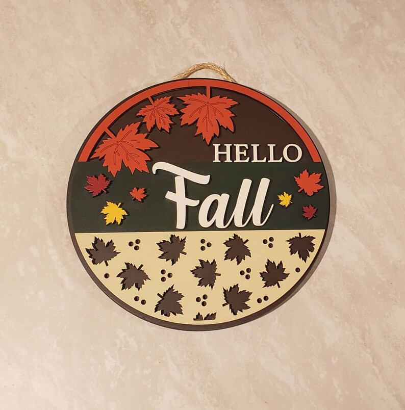 May include: Round wooden sign with the text "Hello Fall" in white lettering. The sign is decorated with red, orange, and yellow autumn leaves and brown leaves on a cream background.
