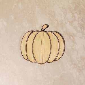 May include: A wooden pumpkin cutout with a natural wood finish. The pumpkin is outlined in black and has a simple, classic design.