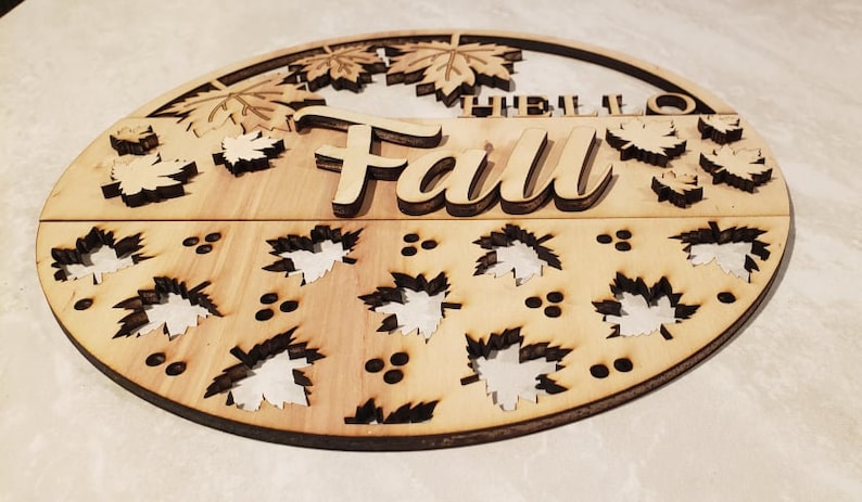 May include: A round wooden sign with the words "Hello Fall" in a decorative font. The sign is decorated with cut-out leaves and berries.