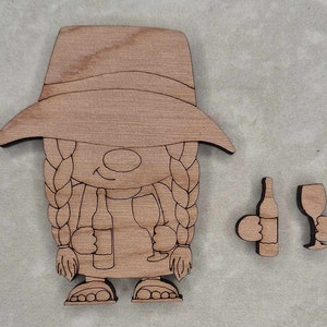 May include: Wooden cutout of a woman wearing a hat and holding a bottle of wine and a glass of wine. The cutout is brown and has a simple design. There are also two separate cutouts of a bottle of wine and a glass of wine.