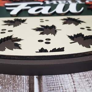 May include: A round wooden sign featuring the words "Hello Fall" in white lettering. The sign is adorned with autumn leaves in a variety of colours and has a brown border.