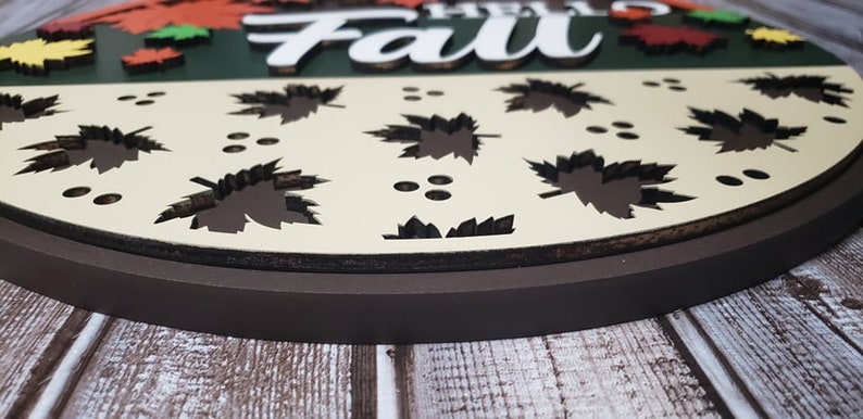 May include: A round wooden sign featuring the words "Hello Fall" in white lettering. The sign is adorned with autumn leaves in a variety of colours and has a brown border.