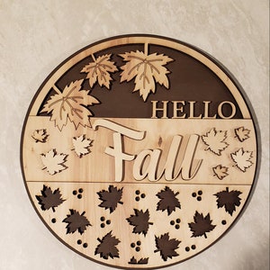 May include: A round wooden sign with the words "Hello Fall" in a brown font. The sign is decorated with cut-out maple leaves in a light brown colour.