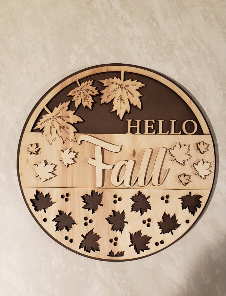 May include: A round wooden sign with the words "Hello Fall" in a brown font. The sign is decorated with cut-out maple leaves in a light brown colour.