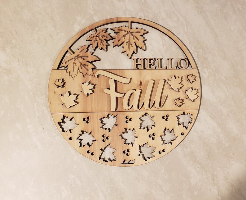 May include: A round wooden sign with the words "Hello Fall" in a decorative font. The sign is decorated with cut-out leaves and berries.