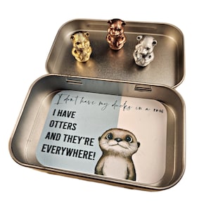 May include: A metal tin with a blue and white label that says "I don't have my ducks in a row I HAVE OTTERS AND THEY'RE EVERYWHERE!" The tin contains three small otter figurines, one gold, one silver, and one bronze.