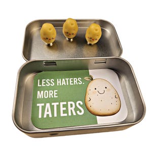 May include: An open, rectangular metal tin with three small, yellow potato-shaped figurines with smiling faces. The tin contains a card with a cartoon potato and the text "LESS HATERS. MORE TATERS."