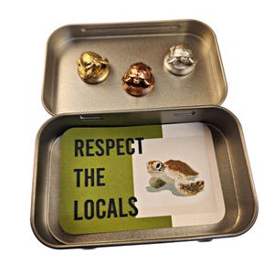 May include: An open, rectangular metal tin with three turtle-shaped pins in gold, copper, and silver. The tin contains a card with a turtle illustration and the text "RESPECT THE LOCALS" in black and white.