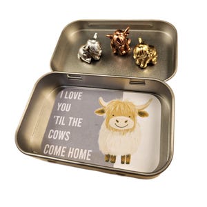 May include: A silver, gold, and copper metal tin with a hinged lid. The tin has a white and gray label with the text "I love you 'til the cows come home". The tin contains three small metal figurines of cows.