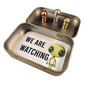 May include: A silver tin with a green alien sticker that says "We are watching". The tin has three small alien figurines inside, one gold, one silver, and one bronze.