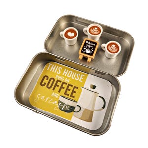 May include: A silver metal tin with a yellow and brown design that says "This house runs on coffee and sarcasm". The tin has a small coffee pot, three miniature coffee cups, and a sign that says "Delicious coffee".