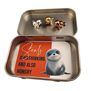 May include: A silver tin with a red and white label that says "Seals Overthinking and Also Hungry". The tin contains three small silver, gold, and copper seal figurines.